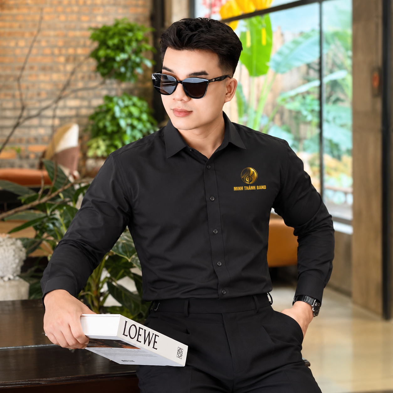 Người mẫu nam mặc áo sơ mi đồng phục đen thêu logo vàng, phối quần tây đen lịch lãm.